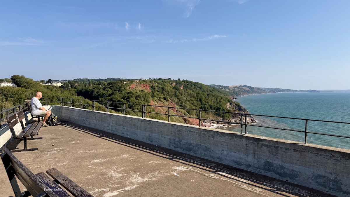 View from the Downs promenade above Oddicombe, with a visitor relaxing on a bench overlooking the Devon coastline.