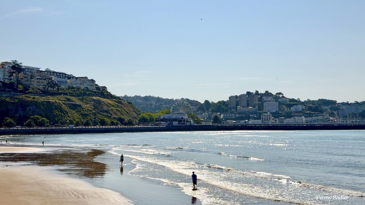 Torre Abbey Sands at low tide, looking toward Torquay's harbour and seafront, with the town's Ferris wheel visible in the distance.