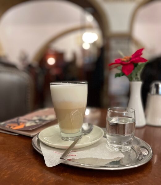 latte coffee on a plate with a spoon and glass of water. A red flower in a white vase is on the table. Cafe Glockenspiel, Linz, Austria.