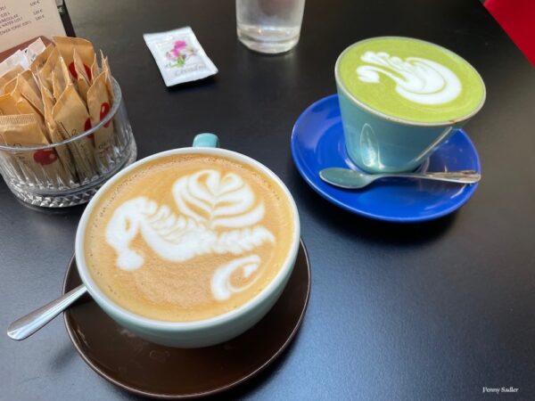 Cappuccino with seahorse foam and matcha latte with swan foam on a black table top with a bowled of sugar packets. Zagreb, Croatia