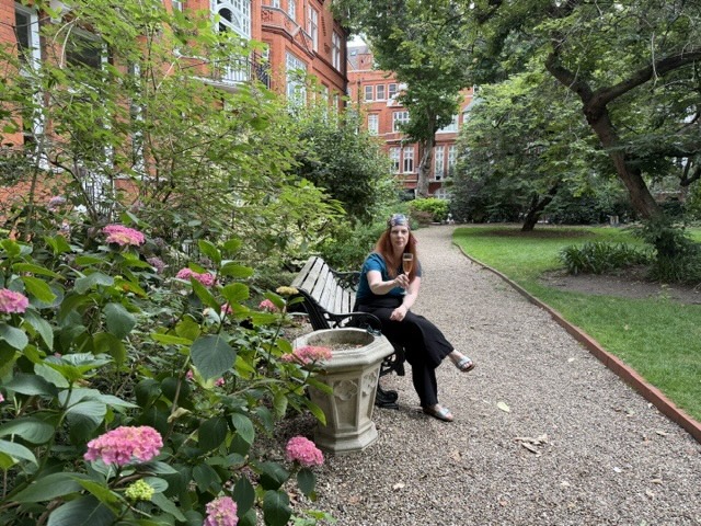 woman holding a glass of sparkling wine sitting in a pretty garden with pink flowers. London