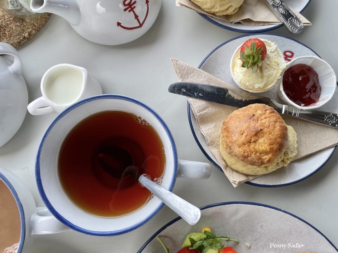 Overhead view of a classic British afternoon tea setting featuring a cup of black tea with a blue rim, a freshly baked scone served with clotted cream, strawberry jam, and a fresh strawberry, alongside a white teapot with a red anchor detail and a small milk pitcher.