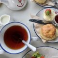 Overhead view of a classic British afternoon tea setting featuring a cup of black tea with a blue rim, a freshly baked scone served with clotted cream, strawberry jam, and a fresh strawberry, alongside a white teapot with a red anchor detail and a small milk pitcher.