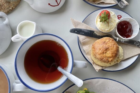 Overhead view of a classic British afternoon tea setting featuring a cup of black tea with a blue rim, a freshly baked scone served with clotted cream, strawberry jam, and a fresh strawberry, alongside a white teapot with a red anchor detail and a small milk pitcher.