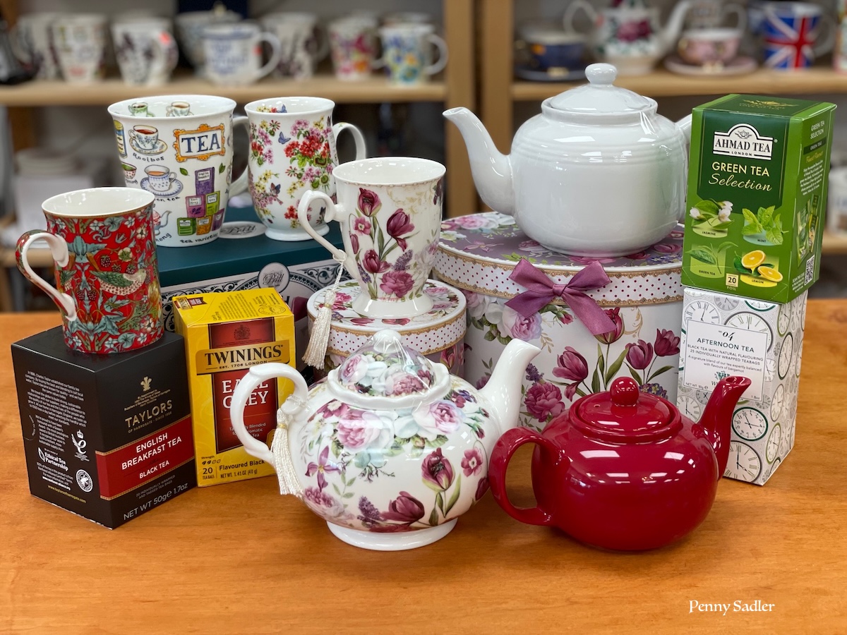 A colorful collection of teapots, tea mugs, and tea boxes including Twinings Earl Grey, Taylors English Breakfast, and Ahmad Green Tea, arranged on a wooden surface with a floral gift box and Union Jack visible in the background.
